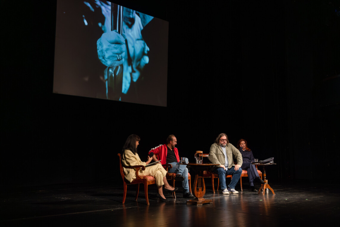 Na scenie przy dwóch oddzielnych, okrągłych stołach siedzą cztery osoby, za nimi na ścianie zawieszony ekran z ciemnym kadrem przedstawiającym dłoń położoną na pionowej rurce do trzymania się w komunikacji