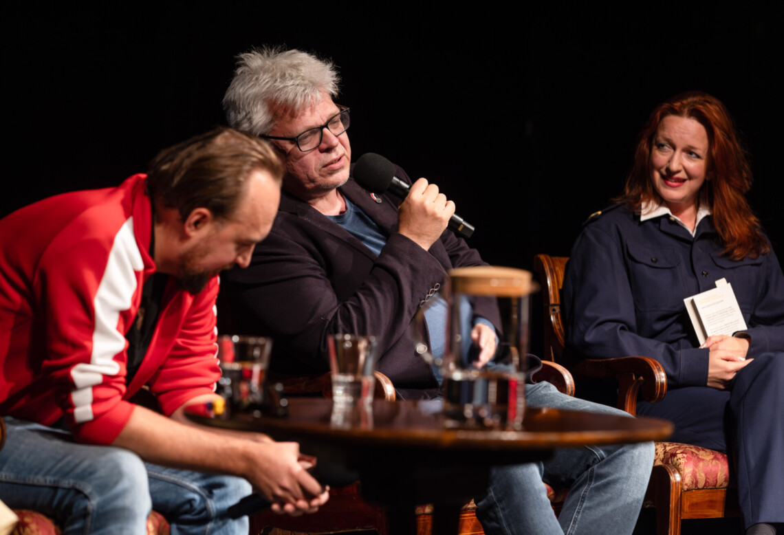 Trzy osoby siedzące na krzesłach, do mikrofonu przemawia siedzący w środku Paweł Czado