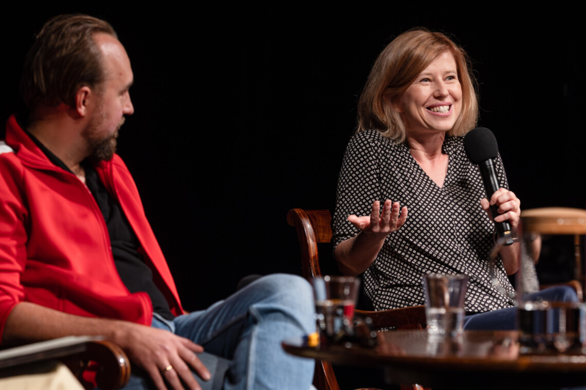 Dwie siedzące osoby, z lewej ujęty z boku Zbigniew Rokuta, obok Bogna Rosińska, która w lewej dłoni trzyma mikrofon, prawą ma skierowaną ku górze, uśmiecha się szeroko odsłaniając zęby