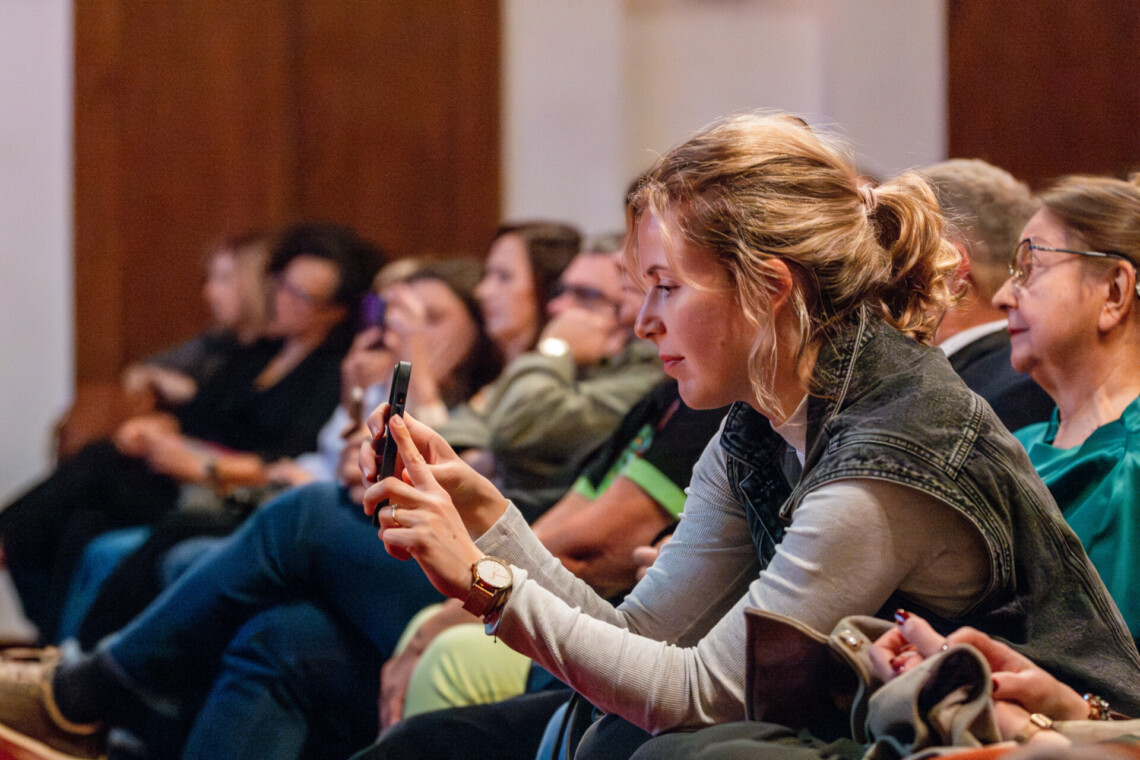 Osoby siedzące na widowni, na pierwszym planie kobieta robiąca zdjęcie telefonem, ujęcie z boku