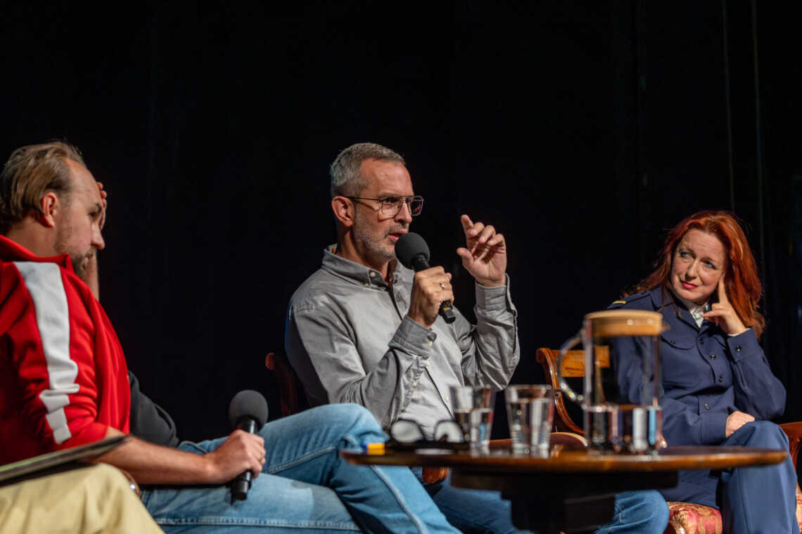 Trzy osoby siedzące na krzesłach, do mikrofonu przemawia siedzący w środku Arkadiusza Gola