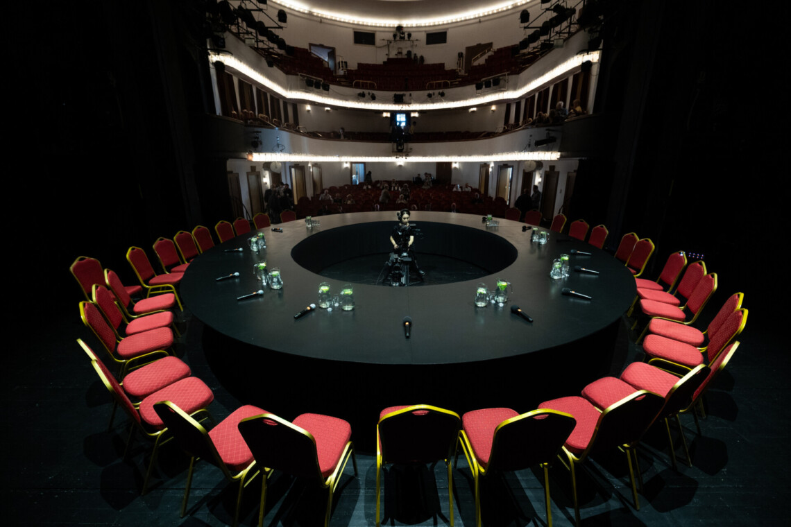 Czarny, okrągły stół wokół którego ustawione są czerwone krzesła, na drugim planie widownia Dużej Sceny Teatru Śląskiego