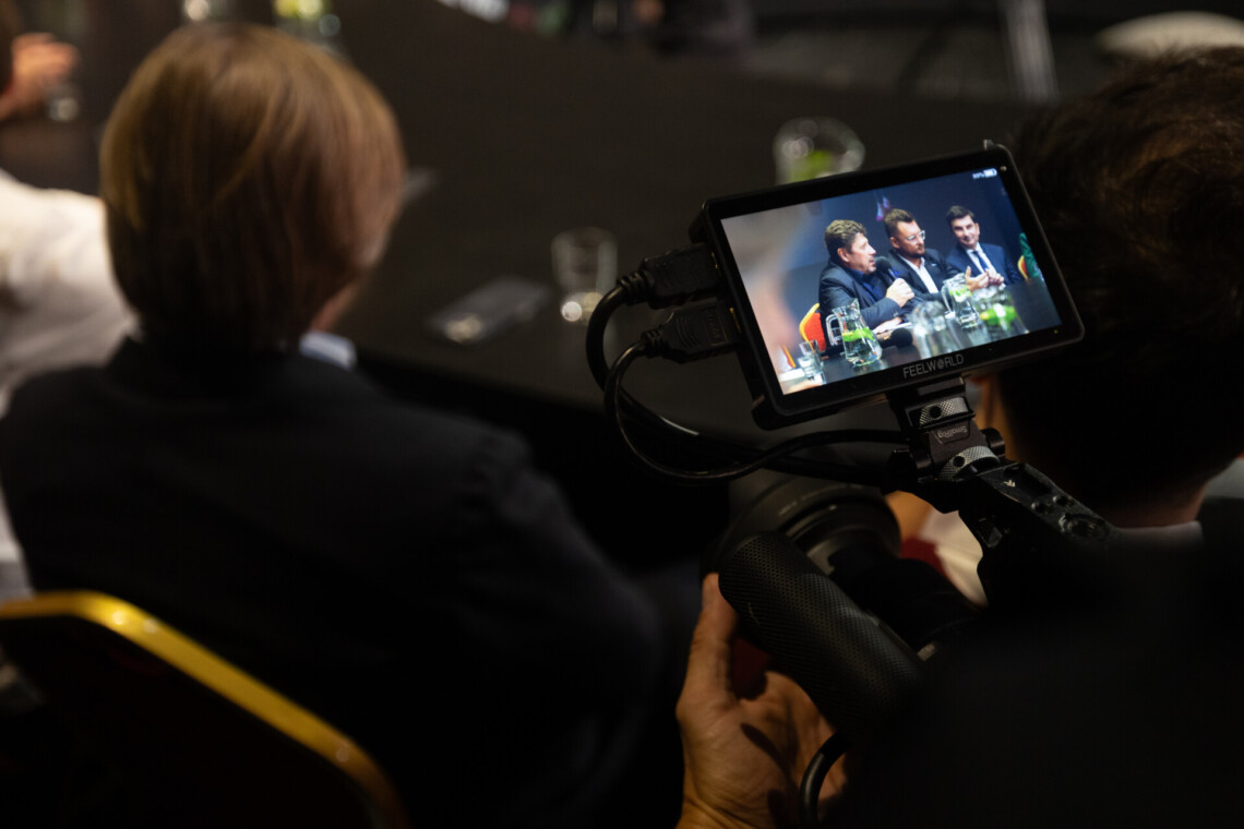 Kamera w trakcie pracy, na dużym wyświetlaczu z podglądem widoczni trzej mężczyźni siedzący obok siebie za stołem