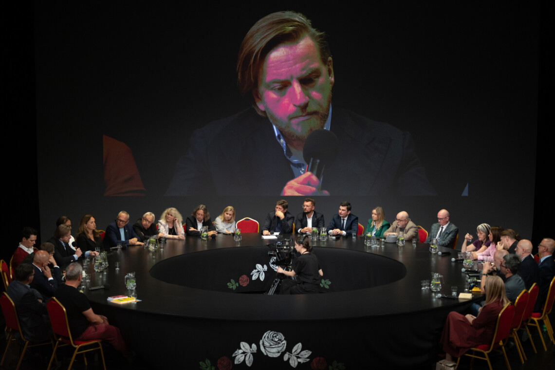 Osoby siedzące wokół czarnego, okrągłego stołu, na ekranie za nimi postać Szczepana Twardocha przemawiającego do mikrofonu