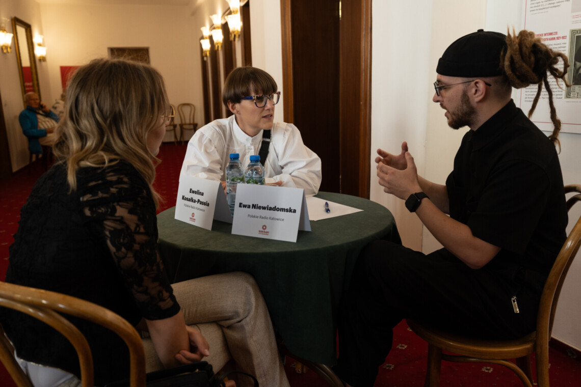 Trzy osoby siedzące przy okrągłym stoliku, na którym stoją dwie kartki z napisami: Ewelina Kosałka-Passia oraz Ewa Niewiadomska