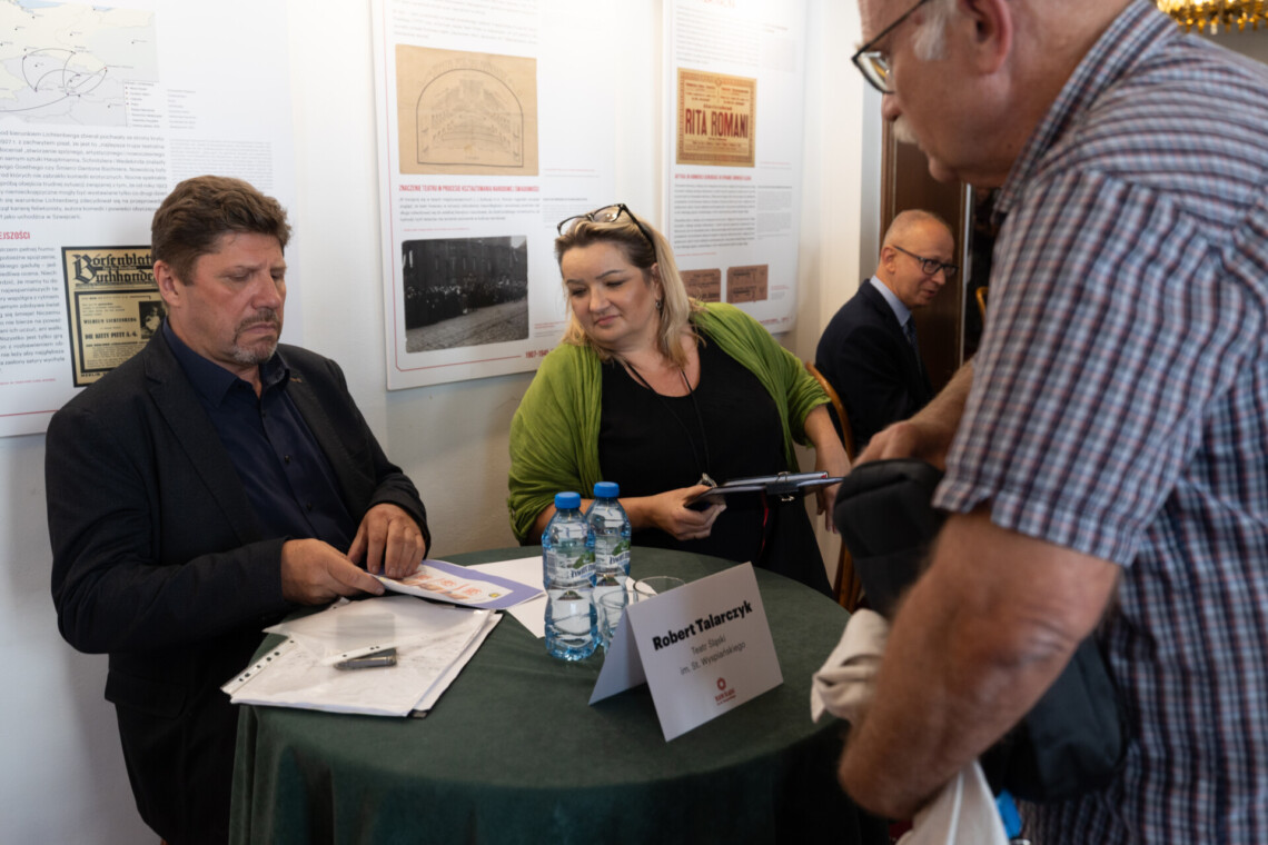 Dyrektor Robert Talarczyk i jego asystentka siedzący przy okrągłym stoliku, na którym stoi kartka z jego imieniem, nazwiskiem i funkcją, patrzą w dół na kartki trzymane przez Dyrektora