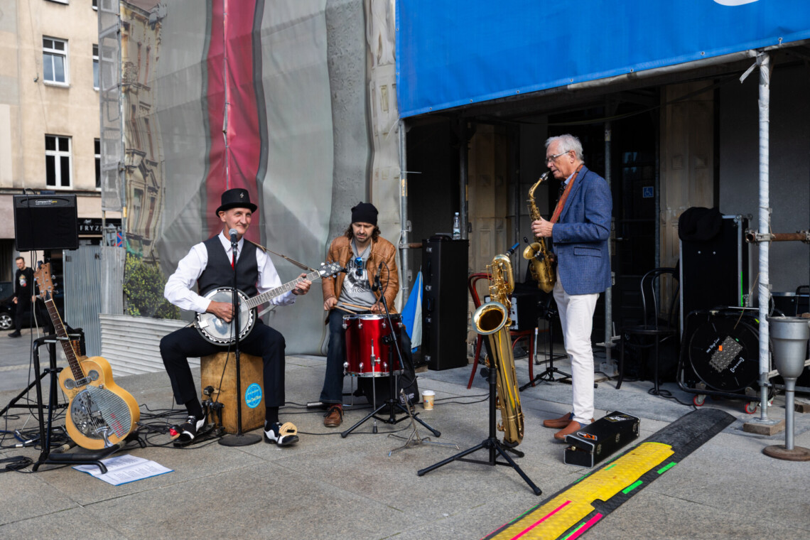 Trzech mężczyzn w trakcie koncertu na zewnątrz, grają na ukulele, bębnie i saksofonie, ujęcie z przodu