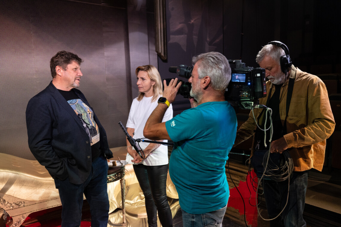 Po lewej stronie dziennikarka przeprowadza wywiad z Dyrektorem Robertem Talarczykiem. Po prawej stronie operator kamer w trakcie nagrywanie, oraz operator dźwięku z mikrofonem na wysięgniku