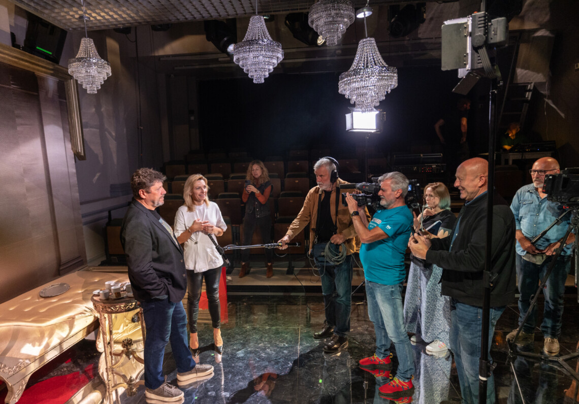 Po lewej stronie dziennikarka przeprowadza wywiad z Dyrektorem Robertem Talarczykiem. Po prawej stronie operatorzy kamer. W tle pusta widownia oraz elementy dekoracji ze spektaklu: kanapa, stolik, kryształowe żyrandole oraz lustrzana podłoga. Ujęcie z oddali