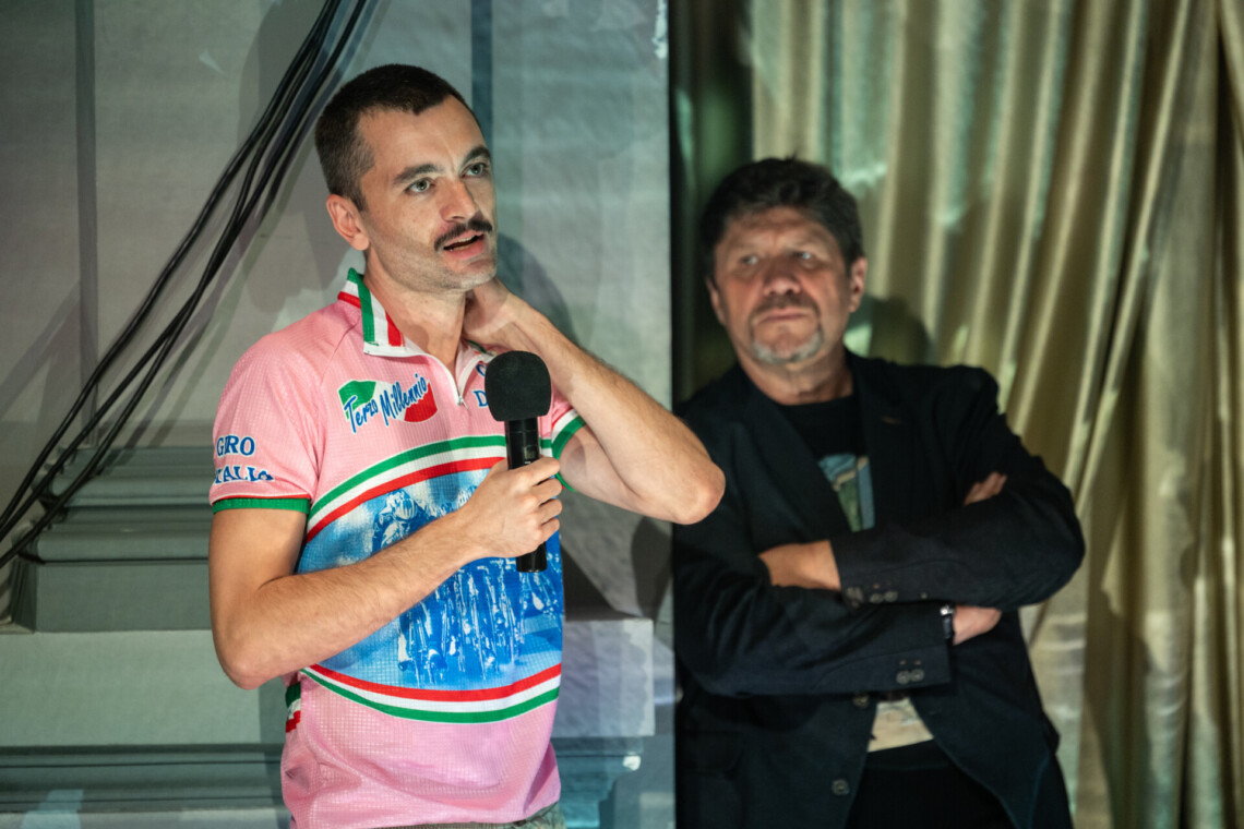 Na pierwszym planie reżyser przemawiający do mikrofonu trzymanego w dłoniach. Na drugim planie Dyrektor Robert Talarczyk, za nim złota kotara.