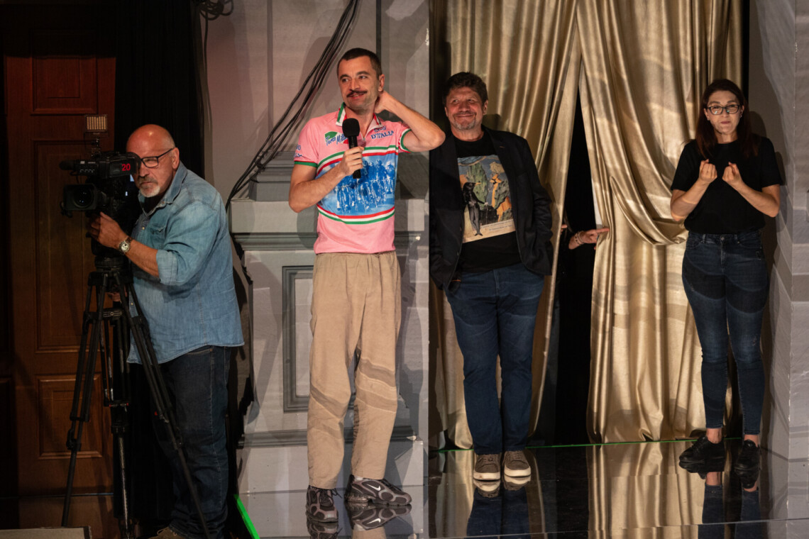 Na scenie cztery osoby. Od lewej kamerzysta, reżyser z mikrofonem w dłoni, Dyrektor Robert Talarczyk oraz Tłumaczka PJM w trakcie pracy. Za nimi złota kotara.