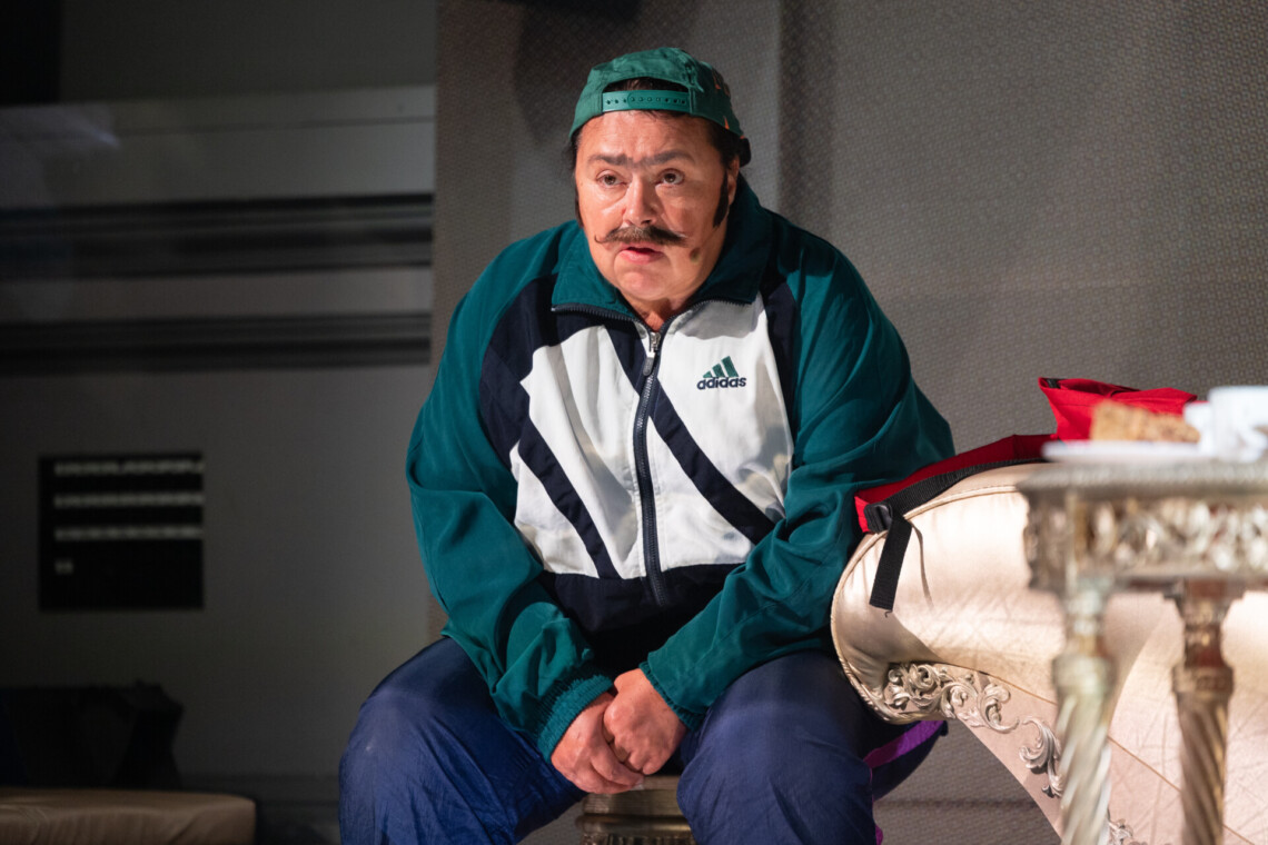 Aktorka stylizowana na mężczyznę siedzi na krześle, jej wzrok skierowany w przestrzeń przed sobą. Postać ubrana w biało-zieloną bluzę dresową oraz granatowe spodnie. Po prawej stronie obok postaci widoczne fragmenty dekoracji: kanapa i stolik.