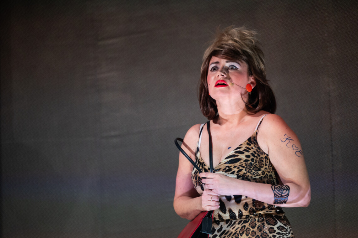 Aktorka w trakcie występu. Kobieta ubrana w sukienkę na ramiączkach w panterkę, włosy ułożone w nieładzie. na lewej ręce namalowane tatuaże. Kobieta w dłoniach ściska pasek z torebki. Tło ciemne.