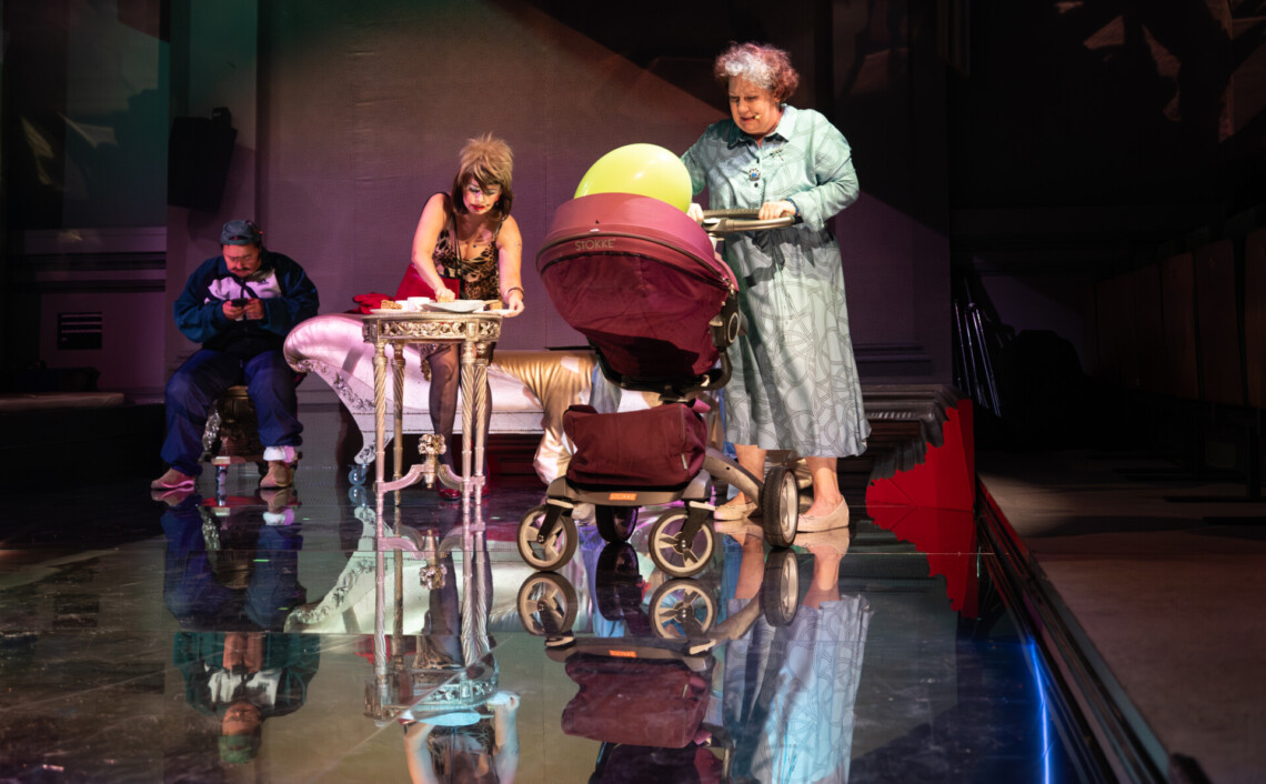 Na pierwszym planie starsza kobieta prowadząca wózek dziecięcy. Na drugim planie kobieta pochyla się nad okrągłym, złotym stolikiem. W tle szezlong i fotel, na którym siedzi mężczyzna piszący na smartfonie. Na podłodze lustrzane kafelki, w której odbijają się występujące postacie.