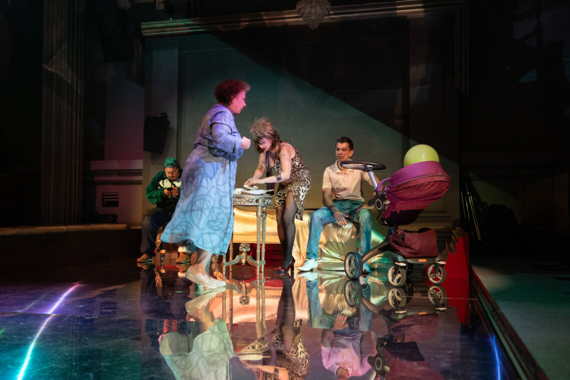 Czwórka aktorów w trakcie występu. Po prawej stronie mężczyzna siedzi na szezlongu w złotym kolorze, spogląda w swoja prawą stronę na kobietę pochylającą się nad stolikiem. Przed i za nimi pozostali aktorzy. Obok pary po lewej stronie stoi wózek dziecięcy w bordowym kolorze. Na podłodze lustrzane kafelki, w której odbijają się występujące postacie.