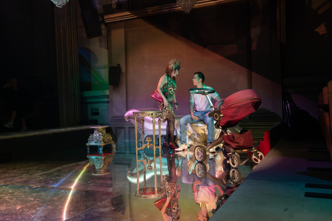 Dwójka aktorów w trakcie występu. Po prawej stronie mężczyzna siedzi na szezlongu w złotym kolorze, spogląda w swoja prawą stronę na kobietę stojącą przed nim. Obok pary po lewej stronie stoi wózek dziecięcy w bordowym kolorze. W tle dekoracja spektaklu. Na podłodze lustrzane kafelki, w której odbijają się występujące postacie.