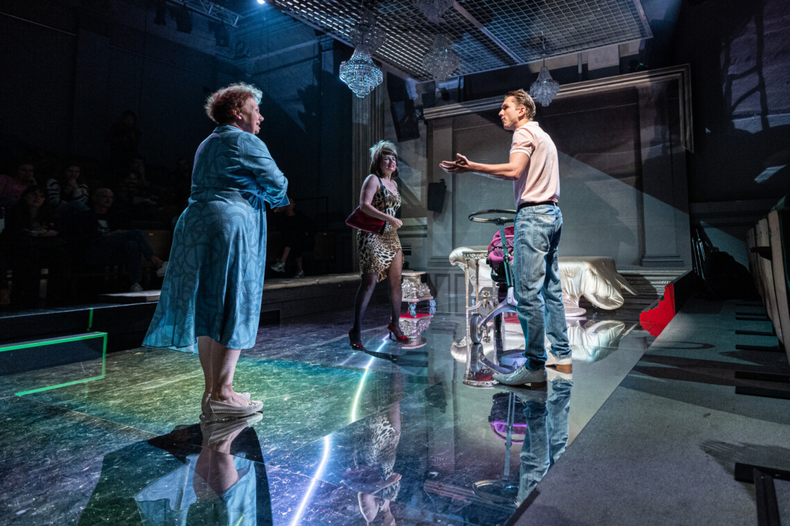 Aktorzy w trakcie występu. Po prawej stronie stoi mężczyzna z uniesionymi dłońmi. Po prawej dwie kobiety kobiety patrzą na mężczyznę. W tle elementy scenografii. Na podłodze lustrzane kafle, w której odbijają się występujące postacie.