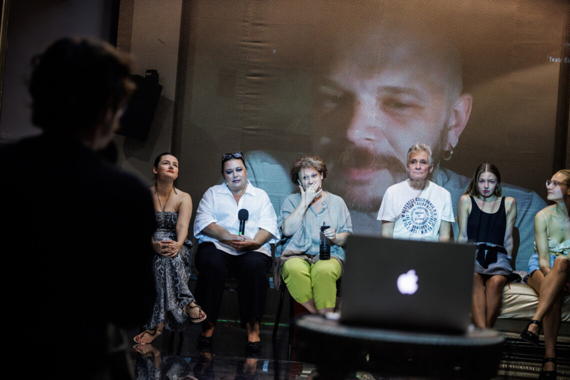 Na pierwszym planie rozmazana postać mężczyzny prowadzącego spotkanie oraz laptop stojący na stoliku, widziane są od tyłu, naprzeciwko siedzą aktorzy, widziani przodem, za nimi na ścianie na ekranie wyświetlony kadr z trwającej rozmowy z reżyserem