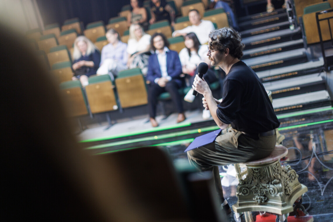 Prowadzący spotkanie mężczyzna siedzi na rzeźbionym taborecie, przemawia do mikrofonu, na drugim planie rozmazane osoby siedzące na widowni