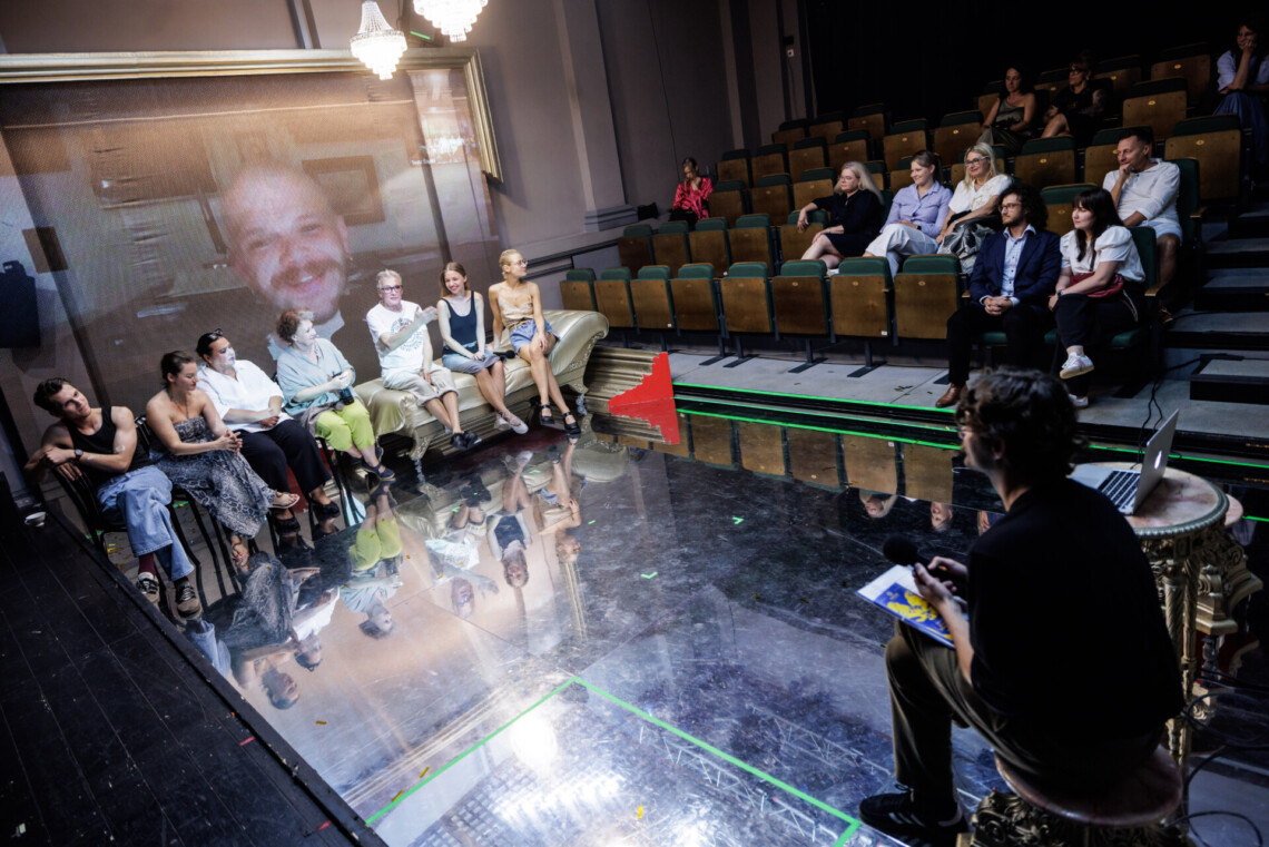 Na scenie naprzeciwko siebie, na przeciwległych końcach siedzą mężczyzna prowadzący spotkanie oraz aktorzy, za aktorami twarz reżysera spektaklu, aktorzy odbijają się w podłodze z luster, z prawej stronie osoby siedzące na widowni