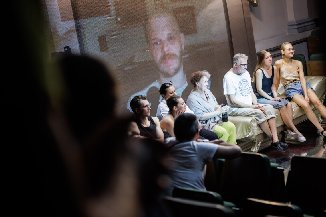 Na scenie w rzędzie siedzą aktorzy, za nimi na ścianie twarz reżysera spektaklu, ujęcie pod kątem z ostatniego rzędu na widowni