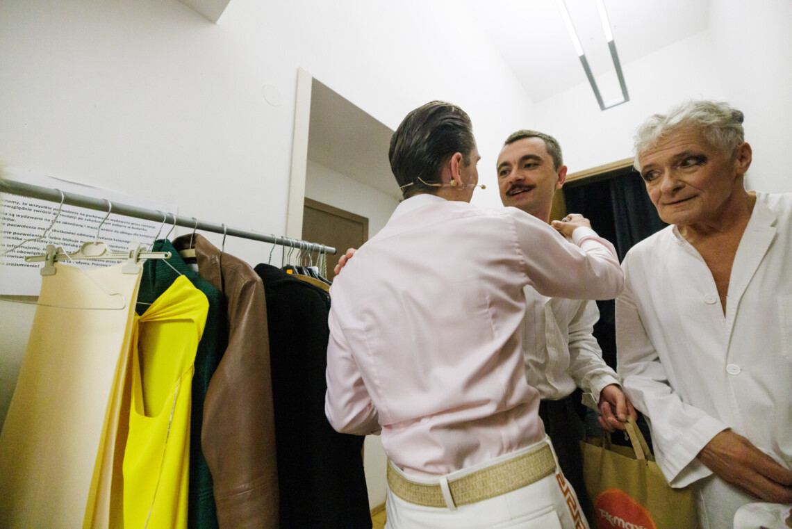 Na pierwszym planie trzech mężczyzn ubranych na biało stoi w korytarzu. Jeden z mężczyzn stoi tyłem do obiektywu, trzyma dłoń na ramieniu jednego z mężczyzn. Na drugim planie podłużny wieszak z zawieszonymi strojami.