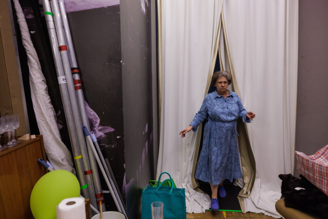 W centrum kobieta przechodząca przez kotarę prowadzącą w kierunku kulis. Ubrana w niebieską sukienkę w geometryczne wzory oraz niebieskie trampki. W tle rekwizyty ze spektaklu.