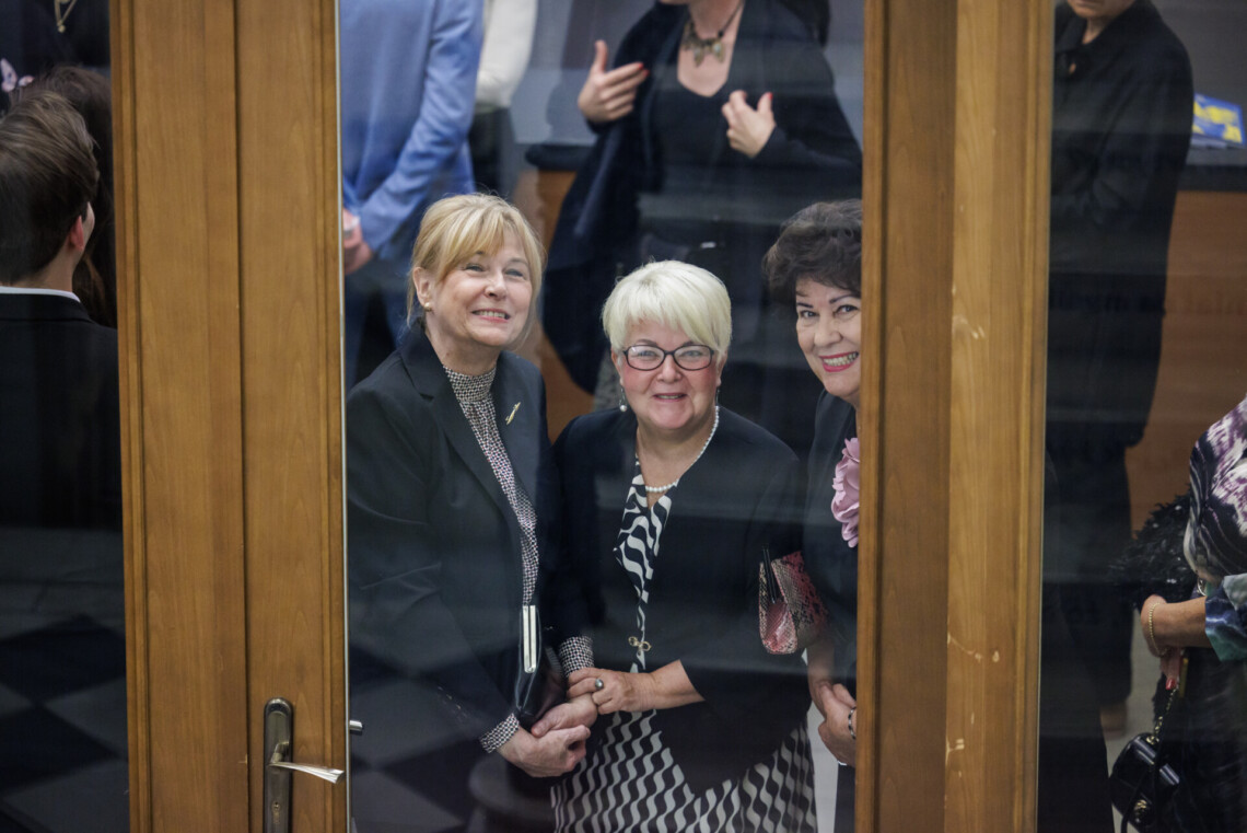 Za zamkniętymi, szklanymi drzwiami stoją trzy uśmiechnięte kobiety. W tle pozostali widzowie.