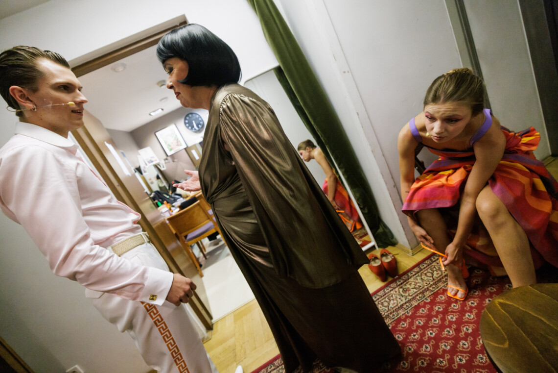 Mężczyzna ubrany na biało stoi na przeciwko kobiety z złotej sukience i czarnych włosach. Po prawej stronie młoda kobieta ubrana w kolorową sukienkę na siedząco zapina sandały. W tle otwarte drzwi prowadzące do garderoby.