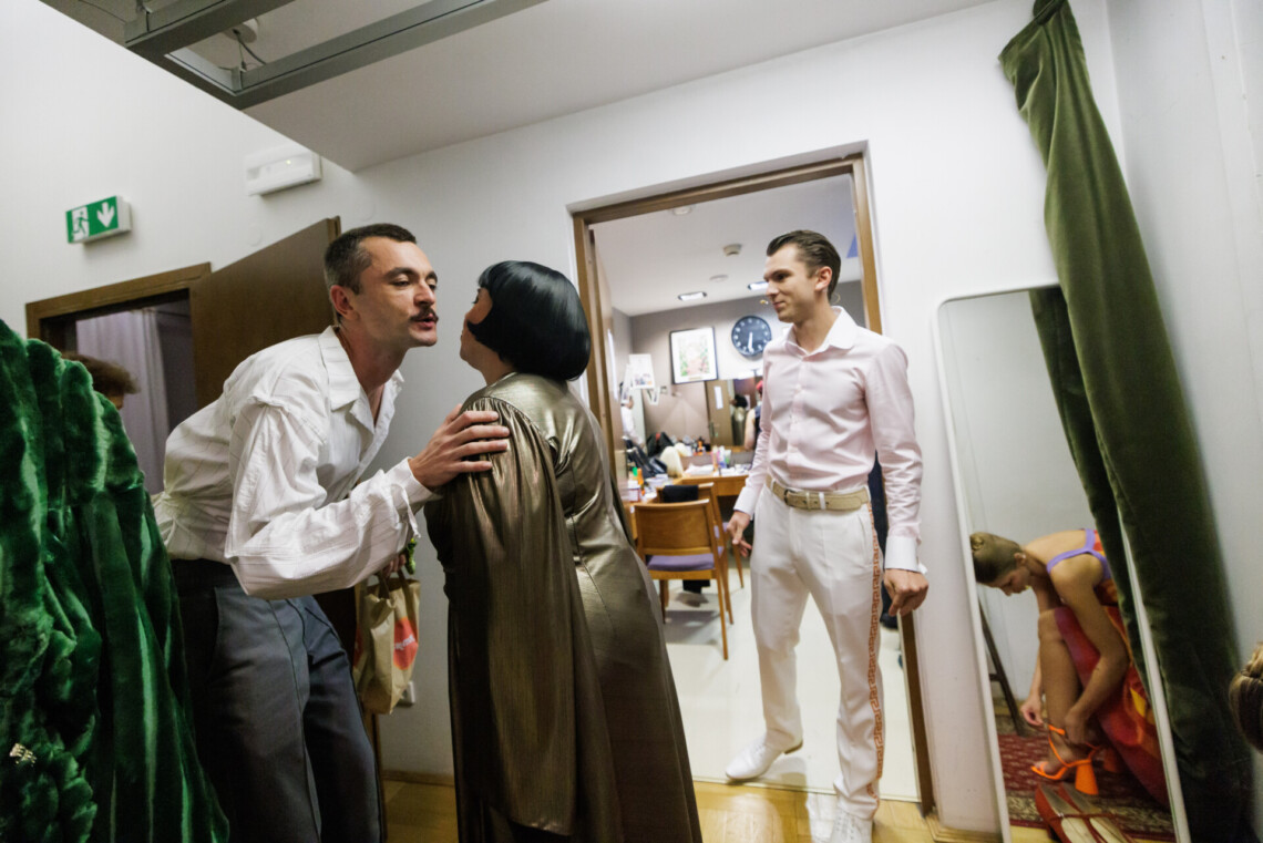 Na pierwszym planie mężczyzna i kobieta całują się w policzek, na drugim planie mężczyzna stojący w w otwartych drzwiach prowadzących do garderoby. Po jego lewej stronie w lustrze odbija się postać kobiety zapinającej sandały.