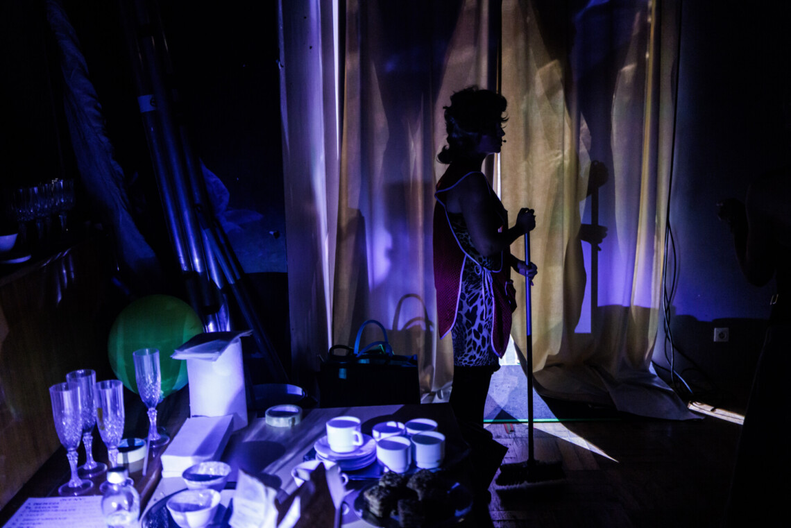 Kobieta stoi w mroku za kulisami, w dłoniach trzyma miotłę. Wokół postaci porozkładane rekwizyty.