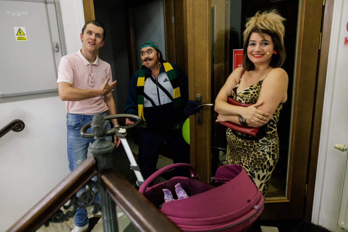 Trójka aktorów w strojach scenicznych stoi przed wejściem na scenę. Przed nimi znajduje się wózek dziecięcy z lalką w środku.