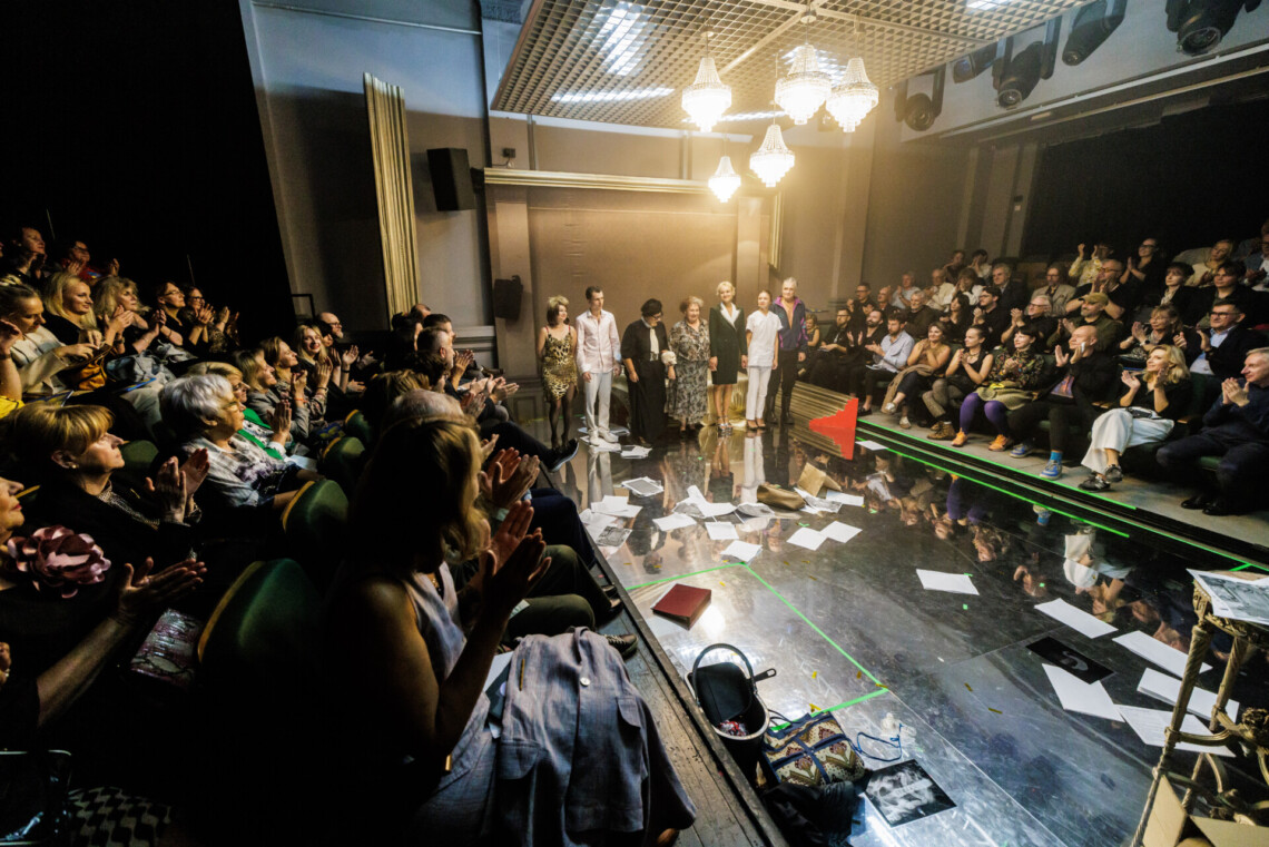 W centrum na scenie w rzędzie stoją aktorzy, z prawej i lewej strony osoby na widowni; ujęcie z oddali