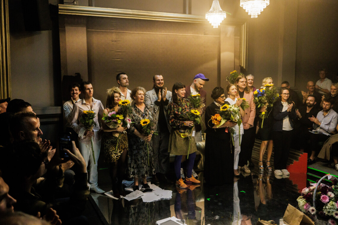 Na scenie w dwóch rzędach z kwiatami w dłoniach stoją aktorzy i realizatorzy spektaklu, z prawej i lewej strony widoczne osoby na widowni; przybliżenie