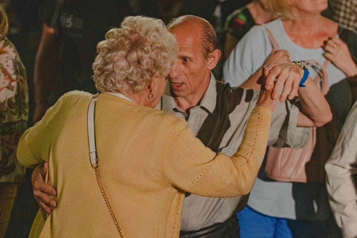 Kobieta i mężczyzna w trakcie tańca w klasycznej pozycji, kobieta odwrócona jest plecami, stoją twarzami naprzeciwko siebie, mężczyzna ma otwarte usta,