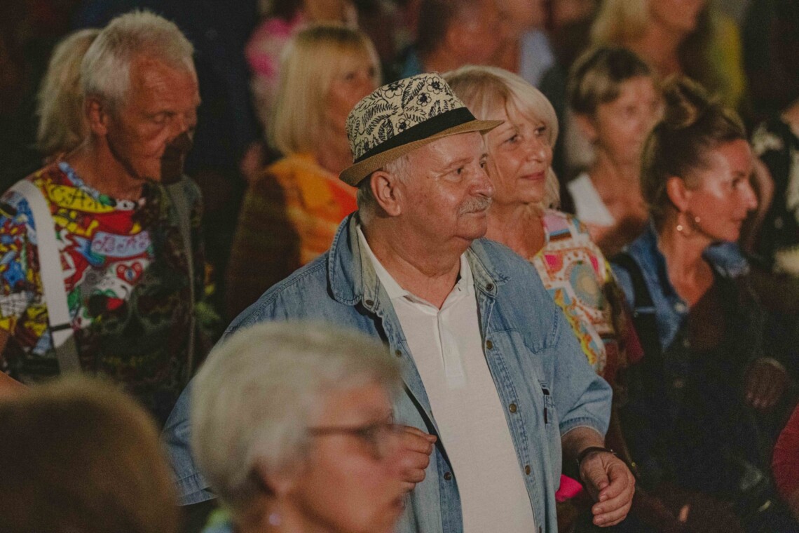 Grupa stojących osób, w centrum starszy mężczyzna w kapeluszu w jeansowej kurtce