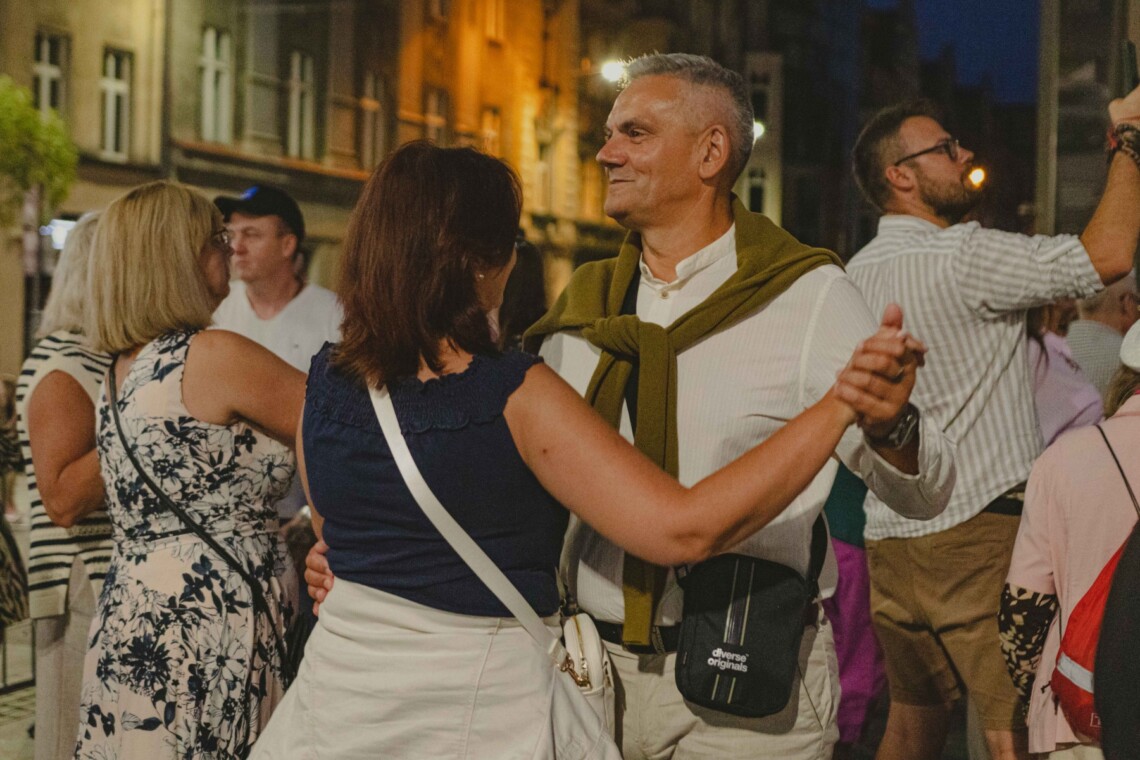 Kobieta i mężczyzna w trakcie tańca w klasycznej pozycji, kobieta odwrócona jest plecami, mężczyzna jest wyższy od niej, za nimi pozostałe tańczące osoby