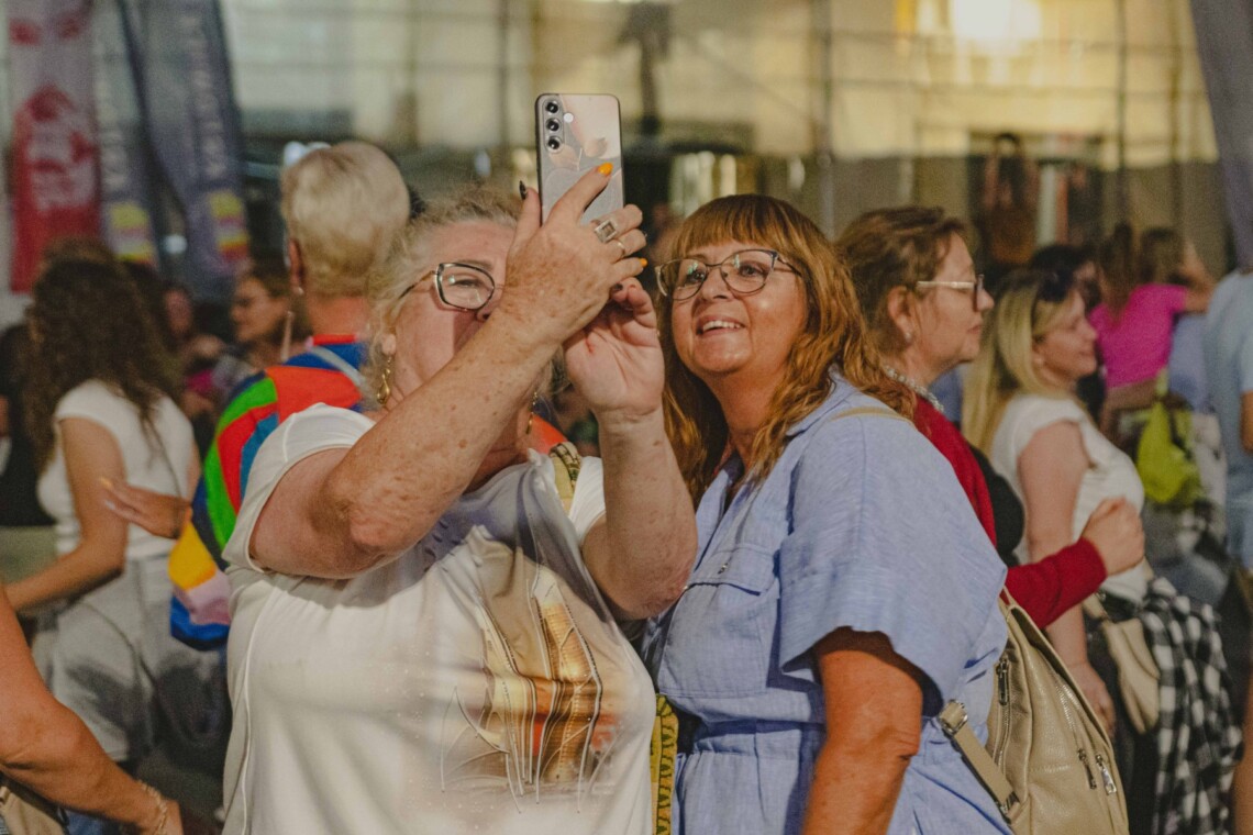 Dwie kobiety stojące razem, jedna z nich robi zdjęcie telefonem, druga patrzy jej przez ramię w telefon