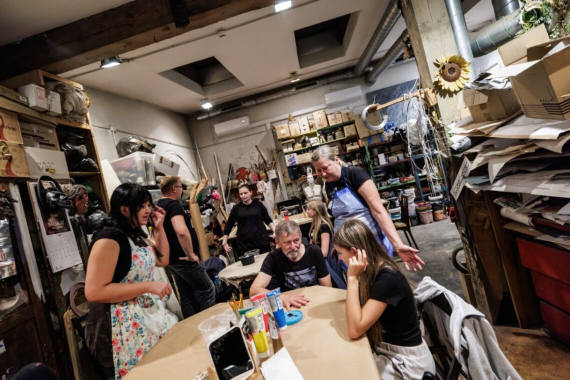 Pomieszczenie pracowni malarskiej. Wokół stołów siedzi lub stoi kilka osób, w tle przedmioty i rekwizyty ze spektakli.