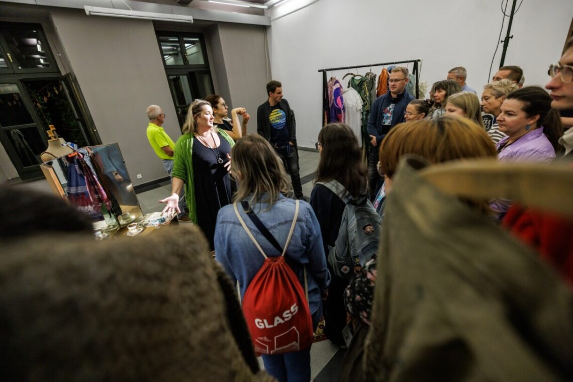 W pomieszczeniu większa grupa ludzi ogląda kostiumy i przedmioty ze spektakli. W centrum kobieta opowiadająca, wokół niej skupieni są zwiedzający