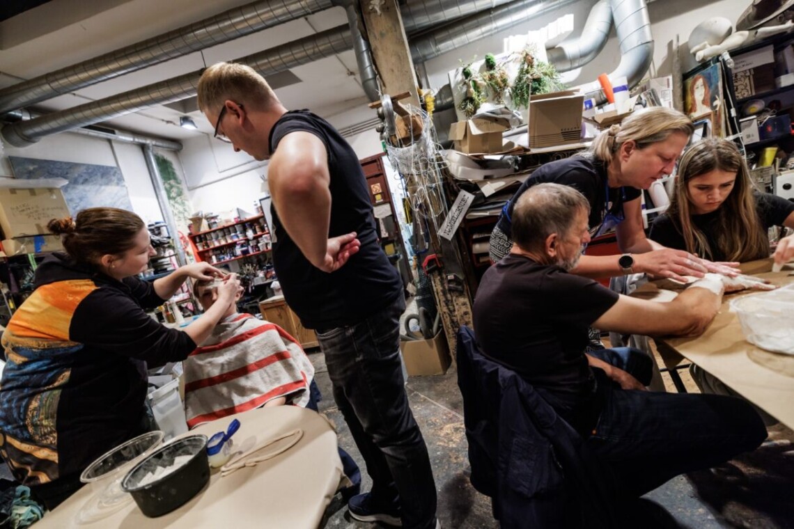 Pomieszczenie pracowni malarskiej. Wokół stołów siedzi lub stoi kilka osób, w tle przedmioty i rekwizyty ze spektakli. Ujęcie z przybliżenia.