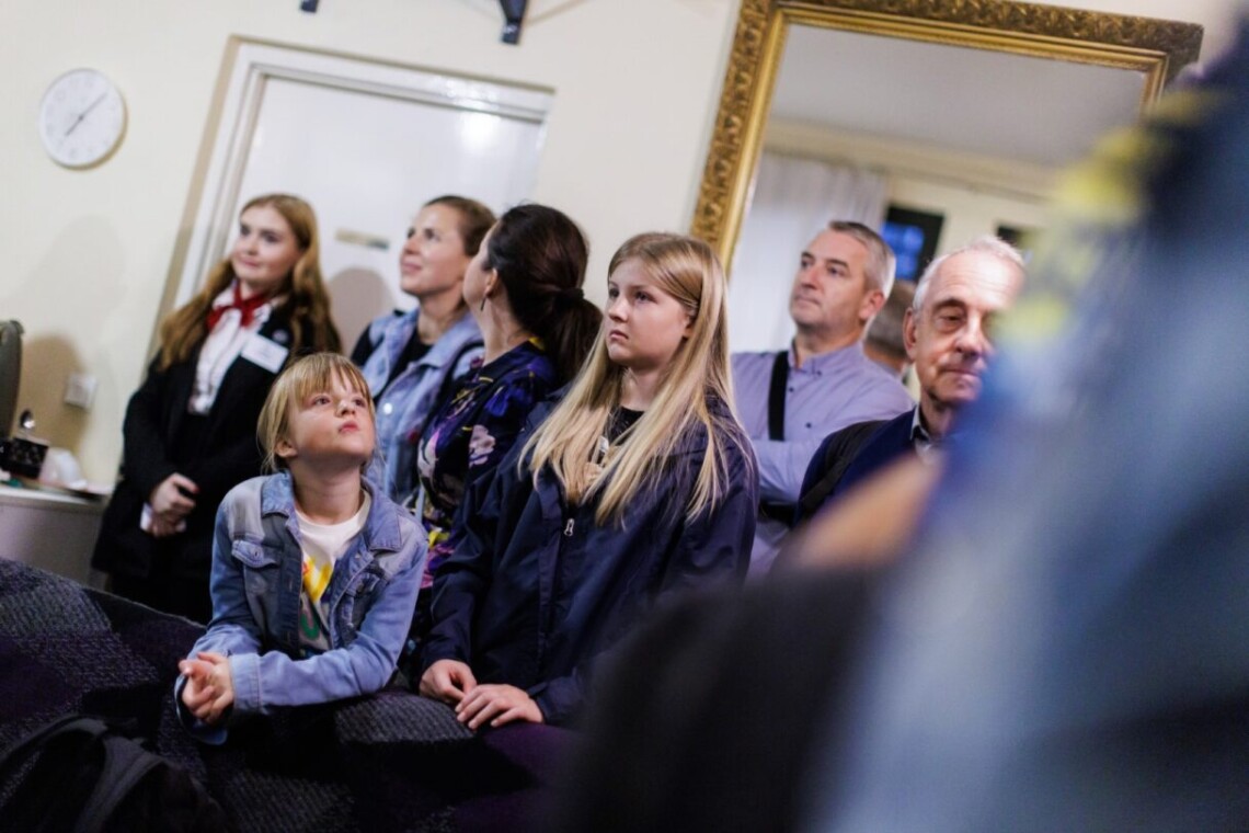 Grupka widzów stoi w zamkniętym pomieszczeniu. Za ich plecami na ścianie wisi duże prostokątne lustro ze złotą, dekorowaną ramą.