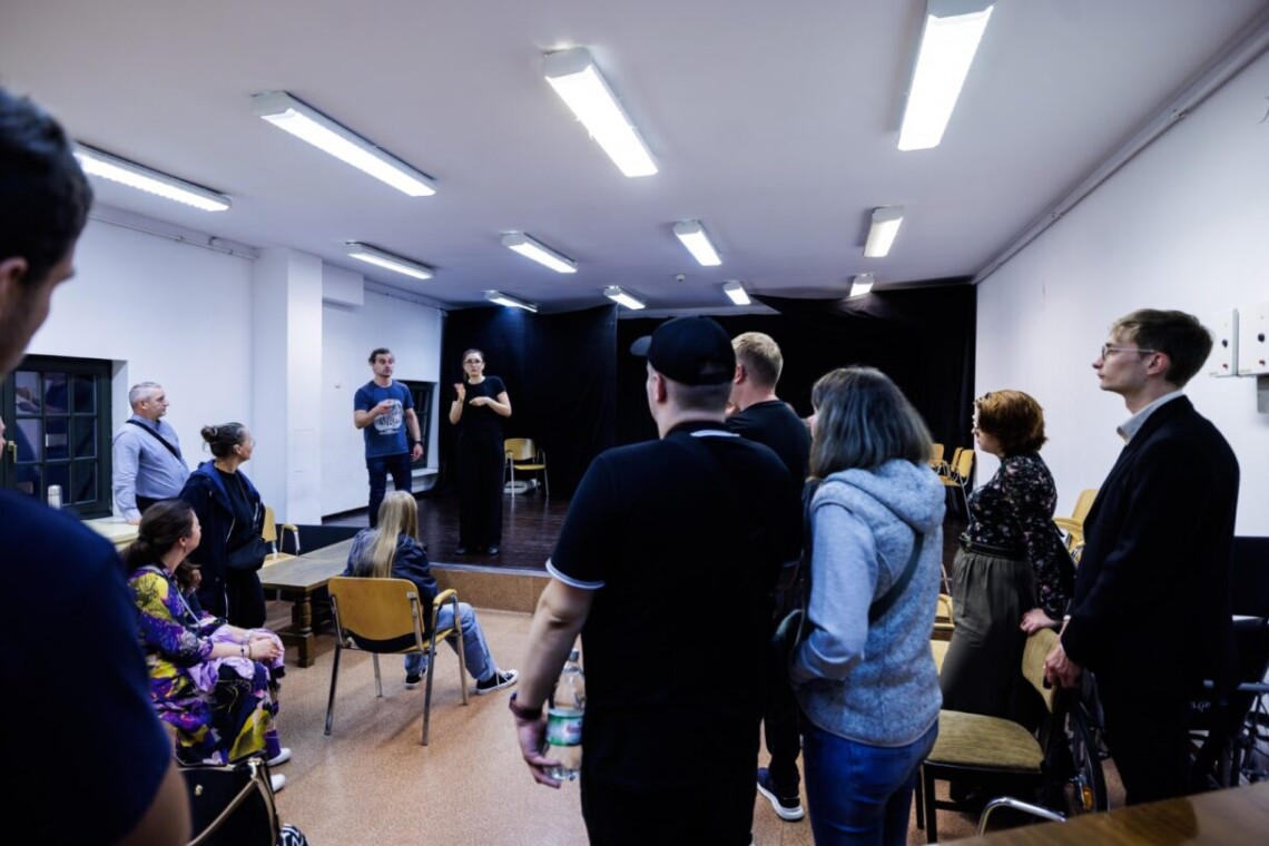 Pomieszczenie przypominające salę prób. Na drugim planie scena z czarnym tłem, na której stoją Bartosz Błaszczyński oraz tłumaczka PJM. Przed nimi poniżej sceny znajdują się zwiedzający.