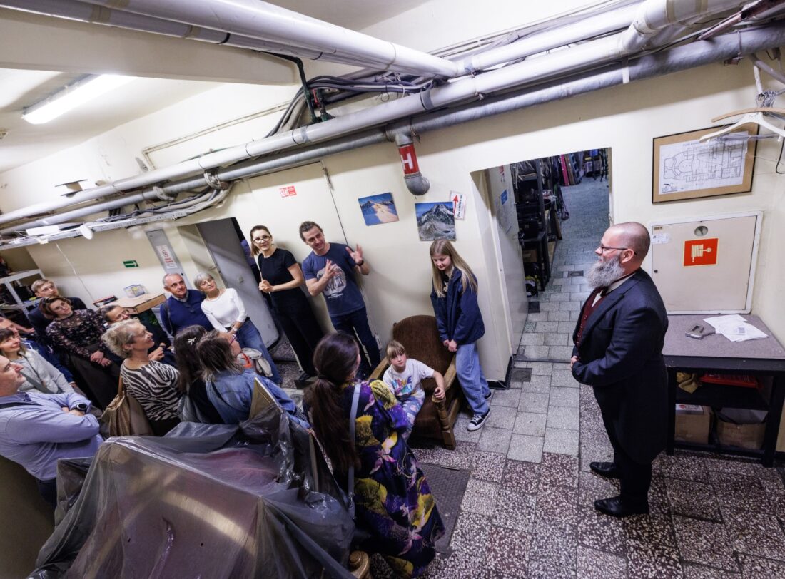 Korytarz w podziemiach Teatru. Po prawej stronie stoi łysy mężczyzna ze szpakowatą brodą, ubrany w granatowy frak. Po lewej stronie stoi grupka zwiedzających, w śród nich aktor Bartosz Błaszczyński oraz Tłumaczka PJM.