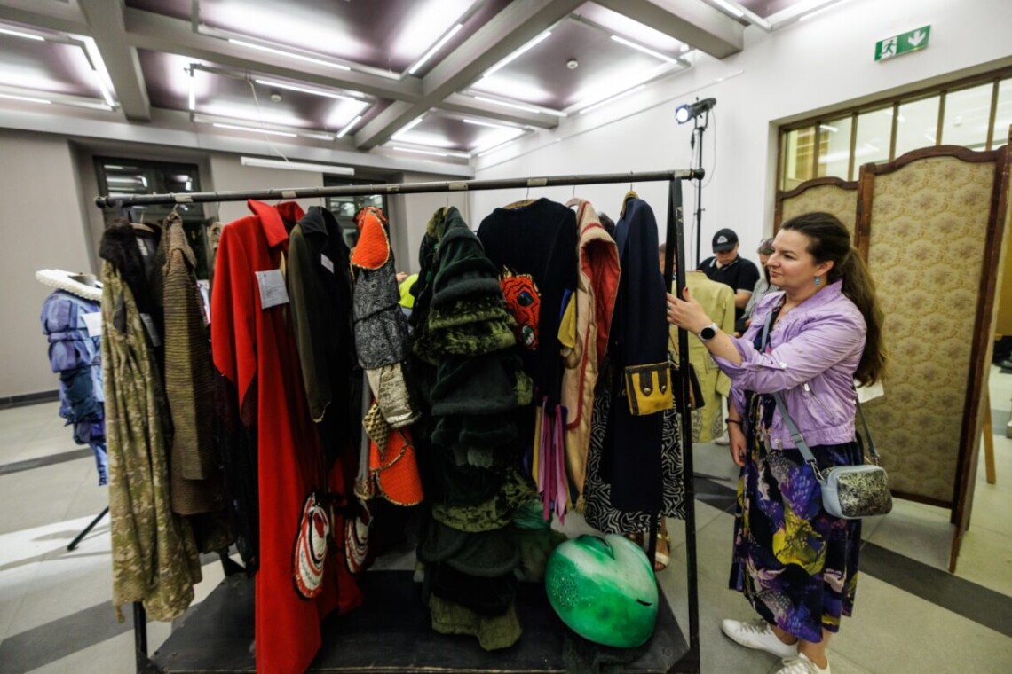W centrum podłużny wieszak z zawieszonymi kostiumami, obok stoi kobieta przeglądającą stroje.