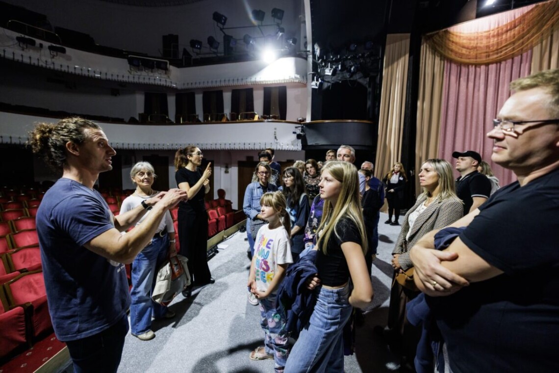 Na pierwszym planie scena duża, na której stoją zwiedzający. Ich wzrok skierowany jest na Bartosza Błaszczyńskiego oraz Tłumaczkę PJM znajdujących po lewej stronie. Na drugim planie pusta widownia na parterze oraz balkony. W tle elementy scenografii, czyli różowe materiały zwisające niczym kurtyna.