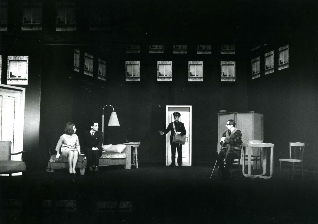 Na scenie przypominającej wysoki pokój czterech aktorów: z lewej strony na kanapie siedzą kobieta i mężczyzna, w centrum przy drzwiach stoi listonosz, z prawej strony na krześle przy stoliku siedzi mężczyzna; zdjęcie czarno-białe, archiwalne