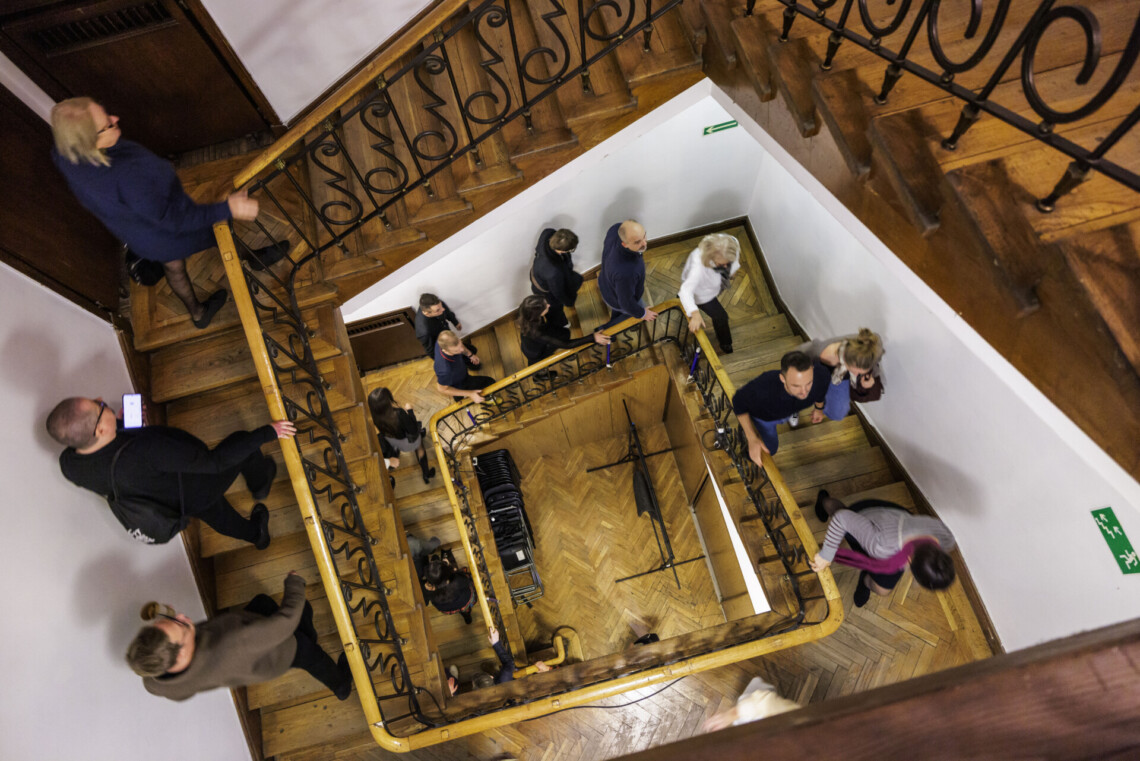 Osoby wchodzące po schodach, ujęcie z góry w dół na trzy piętra klatki schodowej