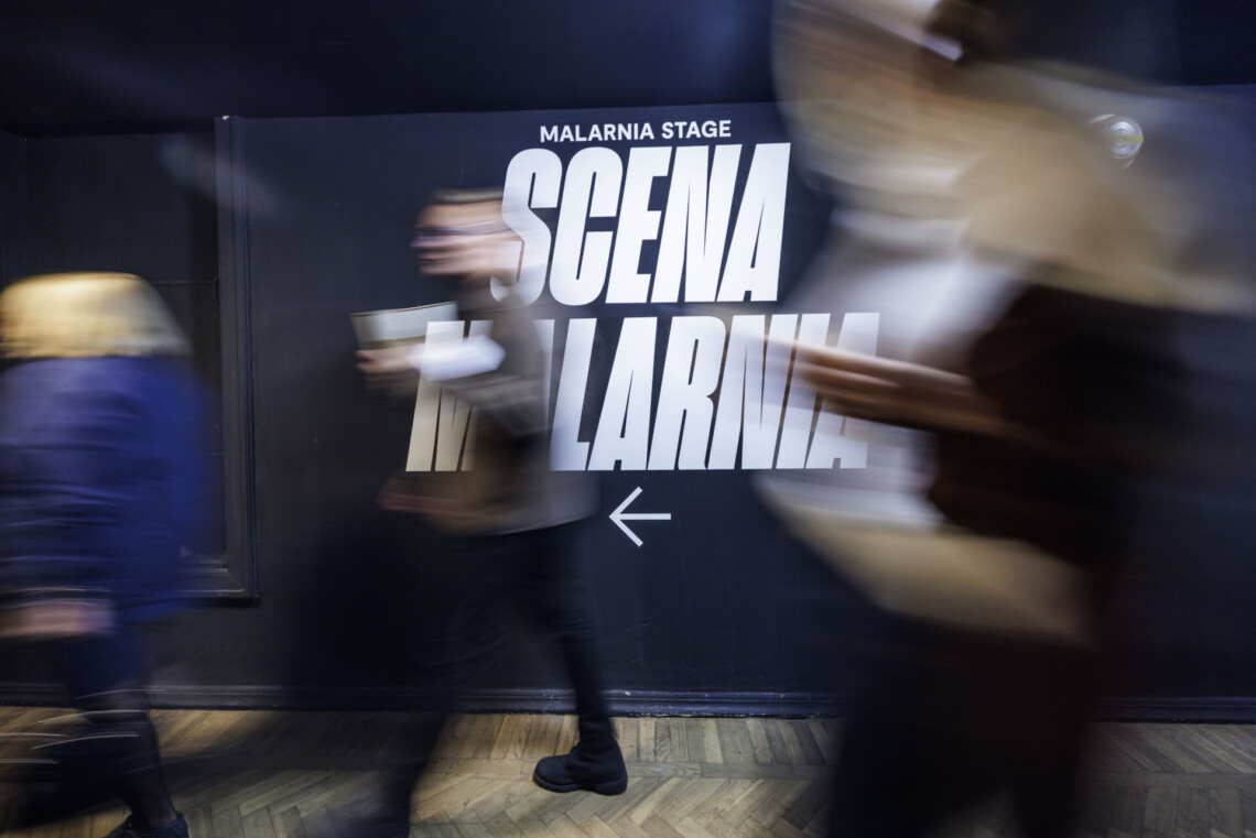 Na ciemnej scenie napis Scena Malarnia, przed ścianą rozmazane kontury przechodzących osób