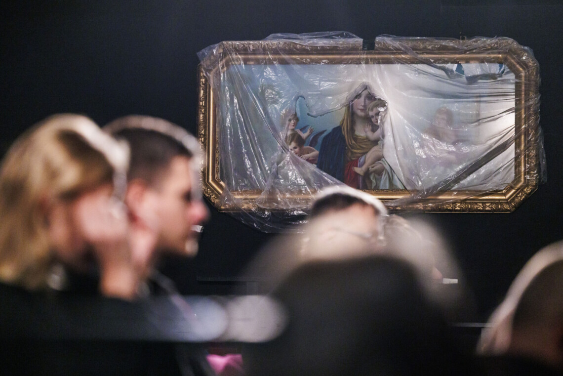 Na ścianie wisi duży, prostokątny obraz Maryi z Jezusem na rękach, obraz częściowo przykryty przezroczystą folią, rozerwaną w środku, na pierwszym planie rozmazane postacie osób siedzących na widowni